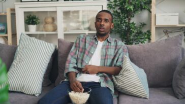 A young man relaxing on a sofa watching TV, holding a remote and popcorn.