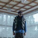 Fashionable man in sunglasses posing indoors wearing a vest and blue sweatshirt.