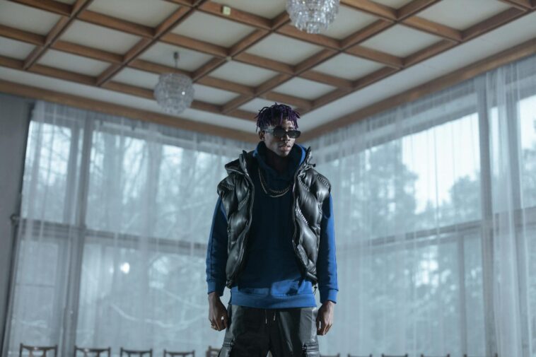 Fashionable man in sunglasses posing indoors wearing a vest and blue sweatshirt.