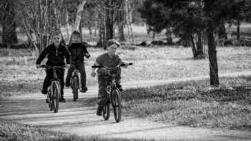 Black and white photo of three kids cycling on a path through a park.