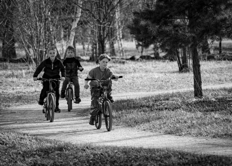 Black and white photo of three kids cycling on a path through a park.