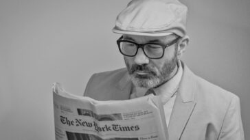 A man in a suit, wearing glasses and a cap, reads The New York Times.