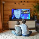 Back view of faceless preteen boy in casual clothes hugging little brother while sitting together on floor and watching TV