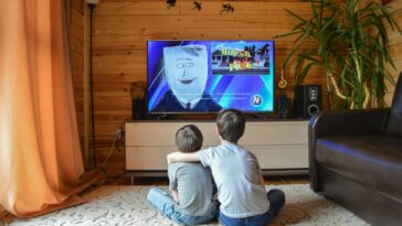 Back view of faceless preteen boy in casual clothes hugging little brother while sitting together on floor and watching TV