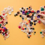 Hand arranging colorful buttons to form a world map on a vibrant yellow background.