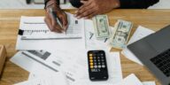 Man working on financial reports with calculator, money, and laptop on a desk.