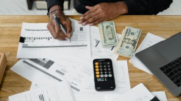 Man working on financial reports with calculator, money, and laptop on a desk.