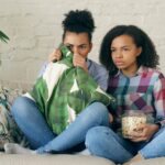 Two young girls sitting on a couch watching a scary movie with popcorn.