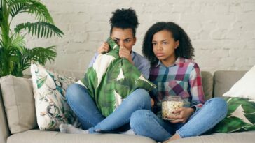 Two young girls sitting on a couch watching a scary movie with popcorn.