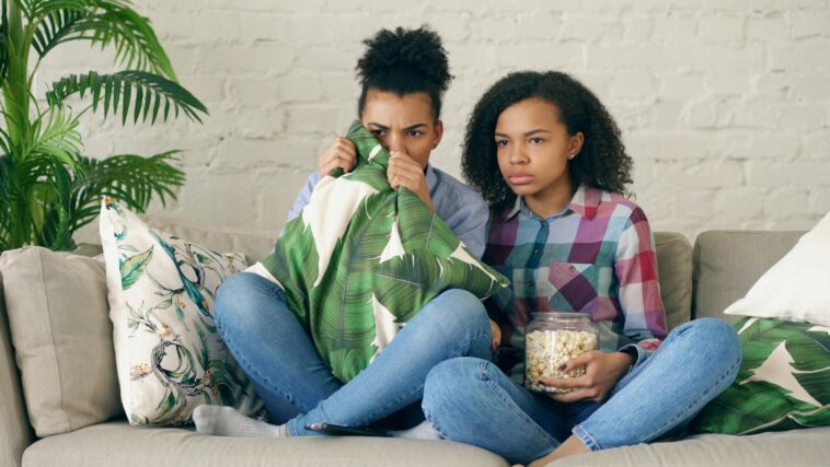 Two young girls sitting on a couch watching a scary movie with popcorn.